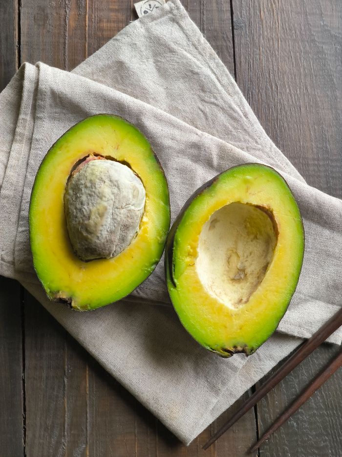 gallery-3 Close-up of a ripe avocado split in half on linen cloth over a wooden surface.